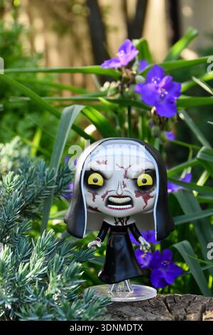 Funko Pop action figure of the Nun from The Conjuring horror movie franchise. Creepy gothic doll in garden among blue flowers on sunny day. Stock Photo
