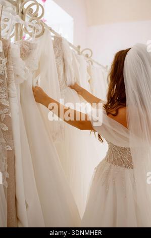 young woman in a white wedding dress and veil looks at wedding gowns displayed in a boutique, seen from behind. She touches and sorts through many wed Stock Photo