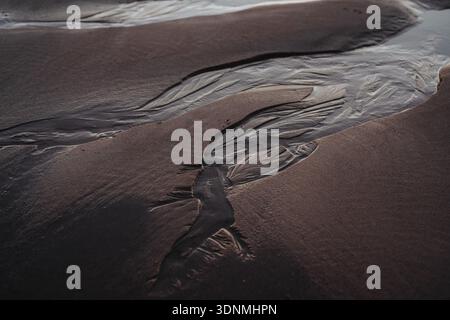 Abstract wet sand patterns with flowing water create sculpted textures on a dark shoreline tonight Stock Photo