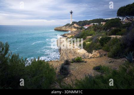 Waves crash against rocky cliffs as the Torredembarra Lighthouse stands tall against a moody sky. Nature thrives along the coast in this scenic spot, Stock Photo