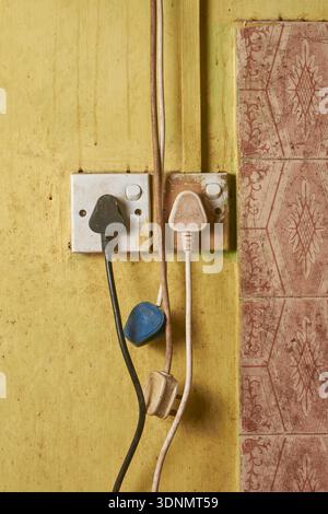 two old plastic electrical sockets with power plugs on weathered yellow wall, disorganized dangling cords with surface grime, electrical hazard Stock Photo