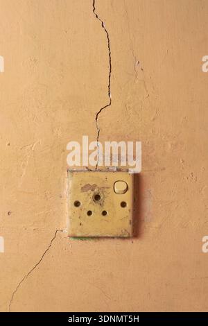 weathered plastic electrical socket on old orange painted wall, vertical crack with surface grime and peeling paint, closeup with copy space, electric Stock Photo
