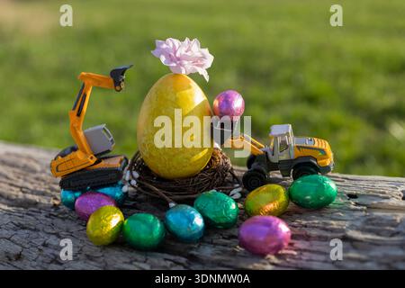 Two toy excavator models, a large painted yellow egg, and lots of colorful chocolate eggs around on a sunny spring day. Business Easter holiday concep Stock Photo