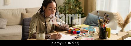 Young woman with braids expresses her creativity while sketching in a stylish room, banner Stock Photo