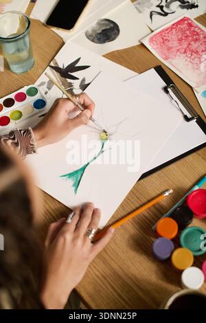 A young woman in stylish clothes paints at her kitchen table full of art supplies. Stock Photo