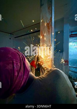 Worker in a pink turban using an angle grinder on a steel beam at a construction site, creating bright yellow sparks in a gritty industrial interior. Stock Photo