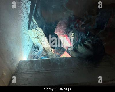 A professional welder in a protective red mask and gloves works in a dark, cramped space, creating bright arcs of light and smoke during fabrication. Stock Photo