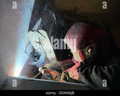 A professional welder in a protective red mask and gloves works in a dark, cramped space, creating bright arcs of light and smoke during fabrication. Stock Photo