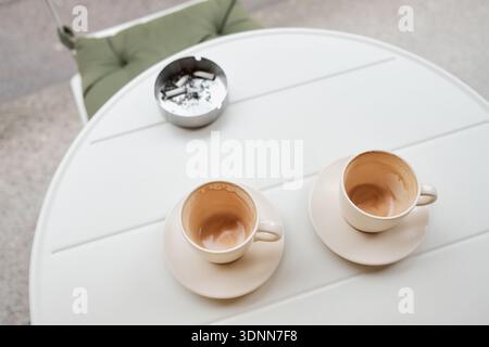 Two empty coffee cups and a metal ashtray with cigarette butts on a white outdoor table. Urban cafe atmosphere showing everyday habits, rest, and Stock Photo