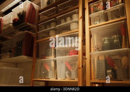 Wooden shelving with transparent plastic containers holding kitchen items, textiles, and decor in a storage room with warm indoor lighting. Stock Photo