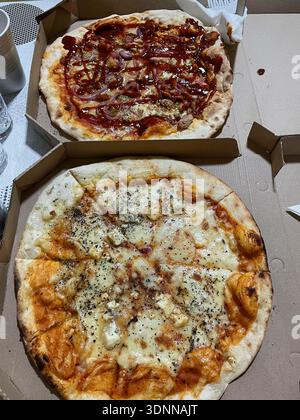 Two freshly baked pizzas served in open cardboard boxes on a table. One pizza topped with meat, onion, and sauce, the other with melted cheese and Stock Photo