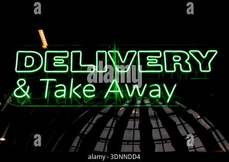 Bright green neon light advertisement for delivery and take away service against a dark background with a subtle architectural reflection below. Stock Photo