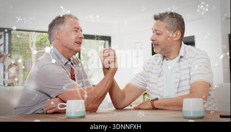 Arm-wrestling two mature men leaning over wooden table at home, clasped hands, orange scarf Stock Photo