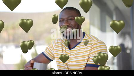 Sitting man holding gray mug on balcony, wearing yellow-striped tee, smiling with metallic hearts Stock Photo