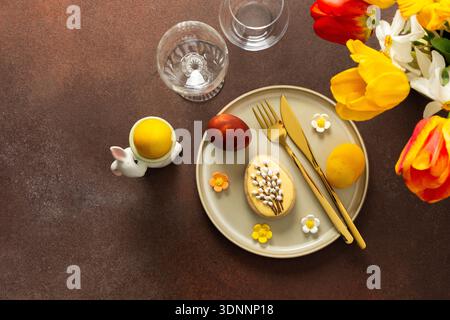 A beautifully arranged Easter table displays vibrant eggs, elegant gold cutlery, and a lively bouquet of tulips and lilies. This setting creates a cheerful atmosphere for the holiday feast Stock Photo