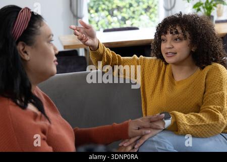 Two women sitting on grey sofa in living room, holding hands, talking by window with wristwatch Stock Photo