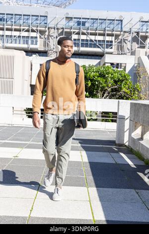 African American male late 20s walking on checkerboard plaza by modern building holding skateboard Stock Photo