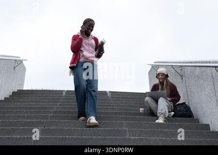 Diverse female friends on campus steps, one using phone holding notes and cup, other using laptop Stock Photo