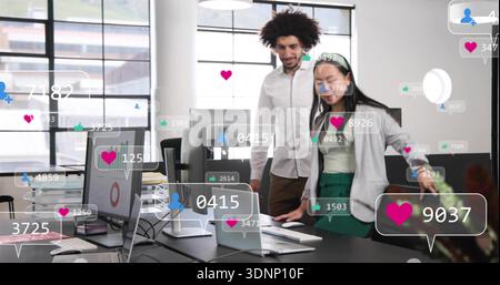 Discussing two colleagues in business wear, leaning over laptop in open office with social icons Stock Photo