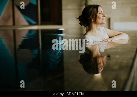 A woman relaxes in a pool at an indoor spa. She sits with her eyes closed and smiles gently. The water reflects her image and the serene environment a Stock Photo