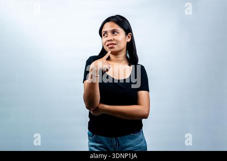 Portrait of a young Indonesian woman with a thoughtful expression, touching her chin and looking up sideways against a white studio background. Stock Photo