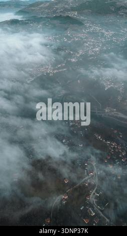 Aerial view of a small mountain village partially covered by low clouds, with houses, fields and forested slopes emerging from fog in soft morning Stock Photo