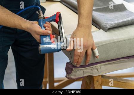 Worker's hand operates a blue pneumatic stapler. Grey patterned fabric is secured on surface. Close-up shows upholstery work, DIY craft, tool use, and Stock Photo