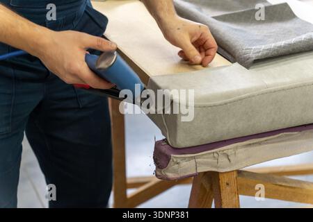 Worker's hand operates a blue pneumatic stapler. Grey patterned fabric is secured on surface. Close-up shows upholstery work, DIY craft, tool use, and Stock Photo