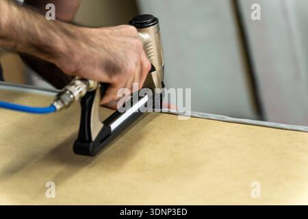 Close-up of upholsterer using a pneumatic staple gun to attach fabric to a wooden furniture frame. Professional workshop repair and DIY renovation con Stock Photo