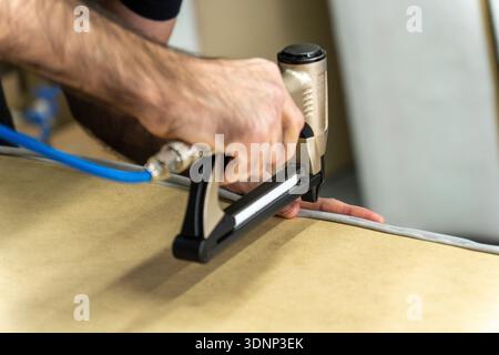 Close-up of upholsterer using a pneumatic staple gun to attach fabric to a wooden furniture frame. Professional workshop repair and DIY renovation con Stock Photo