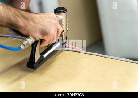 Close-up of upholsterer using a pneumatic staple gun to attach fabric to a wooden furniture frame. Professional workshop repair and DIY renovation con Stock Photo
