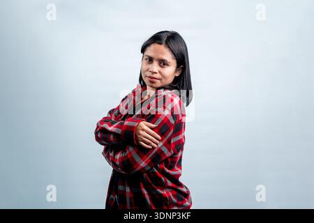 A charming young Southeast Asian woman smiling shyly at the camera, posing with folded arms in a casual red flannel shirt. Stock Photo