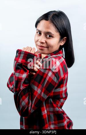 A charming young Southeast Asian woman smiling shyly at the camera, posing with folded arms in a casual red flannel shirt. Stock Photo