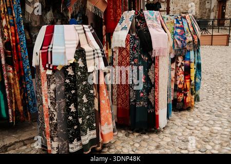 Colorful scarves and shawls hanging at an outdoor market stall on a cobblestone street. Fashion accessories with floral and abstract patterns in a Stock Photo