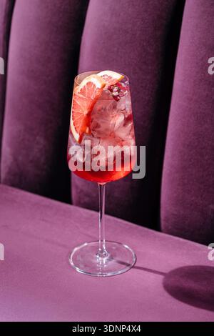 A refreshing grapefruit spritz cocktail garnished with a slice of grapefruit, served in a tall glass with ice, placed on a plush purple velvet chair, Stock Photo