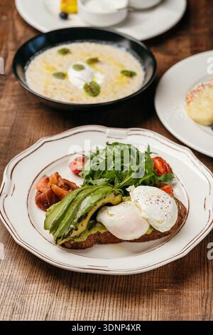 A vibrant meal featuring whole-grain toast topped with creamy guacamole, fresh avocado slices, poached egg, salmon, and arugula, with a bowl of oatmea Stock Photo