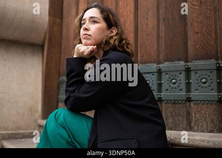 A thoughtful woman seated on outdoor stone steps, rests her chin on her hand. She gazes upward, wearing a black blazer and green trousers against a te Stock Photo