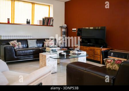 A modern living room featuring leather sofas, a flat-screen TV on a wooden console, and a glass coffee table. The warm lighting and decorative pillows Stock Photo