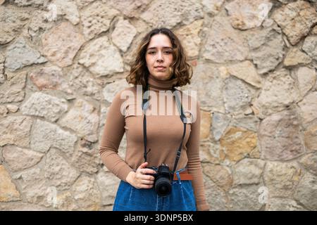 A woman with a casual outfit and a camera stands against a textured stone wall. She wears a brown turtleneck and blue jeans, conveying a relaxed yet p Stock Photo
