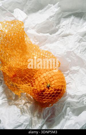 A vibrant citrus fruit is wrapped in orange mesh, resting on a textured sheet of crumpled white paper. Stock Photo