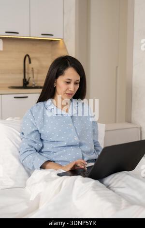 A woman in comfortable pajamas works from her bed using a laptop, conveying a relaxed and cozy home environment. The setting suggests productivity and Stock Photo