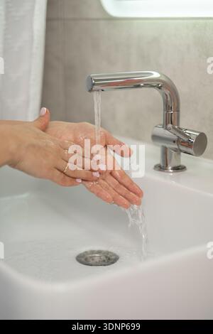 A close-up image of a woman washing her hands under running water in a modern bathroom sink. The focus is on hygiene, self-care, and cleanliness in a Stock Photo