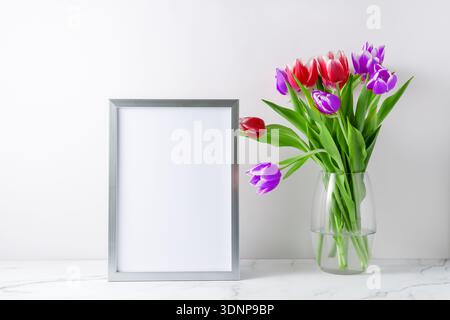 Bouquet blooming red purple white tulip flowers in glass vase and empty poster or photo frame mockup on white marble table in interior. Light backgrou Stock Photo