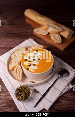 warm autumnal scene featuring creamy pumpkin soup garnished with seeds and cream alongside fresh baguette slices on a rustic wooden table setting, evo Stock Photo