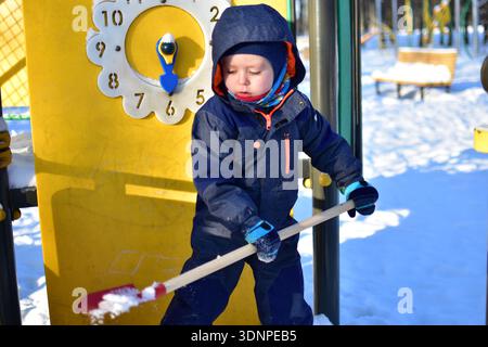 Toddler boy playing with red shovel near yellow playground panel with clock design in snowy park. Child enjoying outdoor winter activity on bright sun Stock Photo