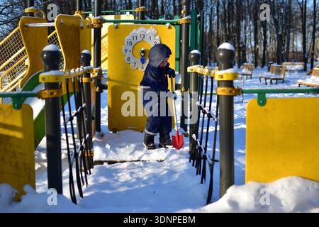 Toddler boy standing on snowy playground bridge holding small red shovel. Child dressed warmly exploring winter park on sunny cold day with snow cover Stock Photo
