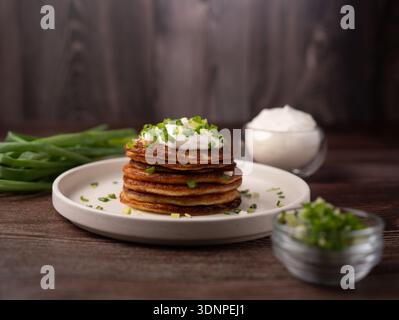 stack of golden potato pancakes topped with a dollop of sour cream and chopped chives on a ceramic plate, arranged on a rustic wooden table with bowls Stock Photo