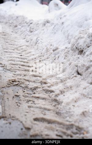 close-up study of slushy plowed snow along a city sidewalk, showing dirty beige-gray ruts, tire tracks and compacted white snowbanks, evoking cold, gr Stock Photo