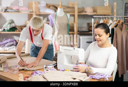 Attentive woman tailor sewing textile using sewing machine and young male assistant drawing pattern on sheet of paper using curves in fashionable Stock Photo