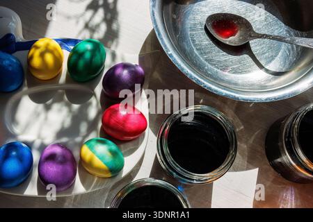 Closeup of brightly dyed eggs on a palette beside glass jars of dark dye and a metal bowl with a stained spoon on a sunlit tabletop. Stock Photo